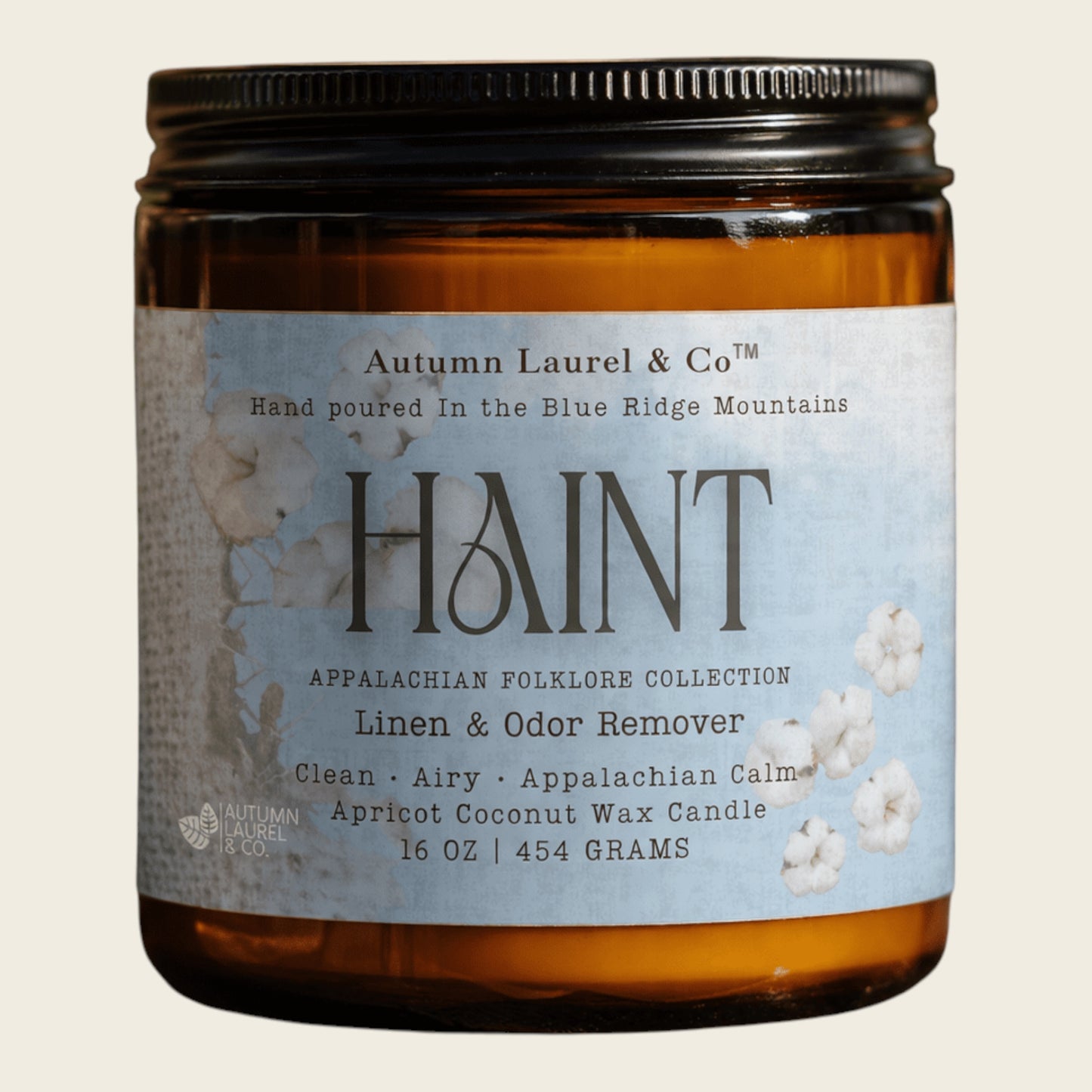 An amber glass jar candle with a black metal lid and a light blue label reading 'HAINT' by Autumn Laurel & Co, photographed on a plain light beige background.