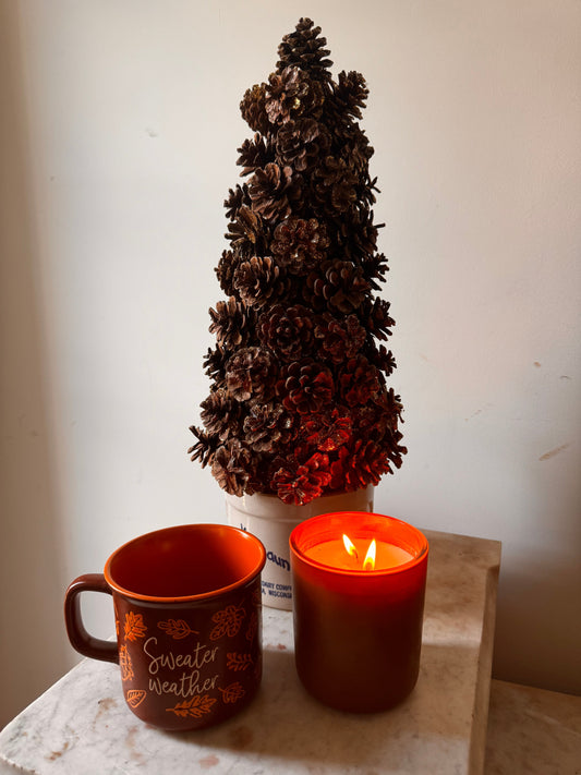 Morning Mercantile Autumn Laurel candle sitting nest to a Fall Coffee Cup and a pinecone tree. 