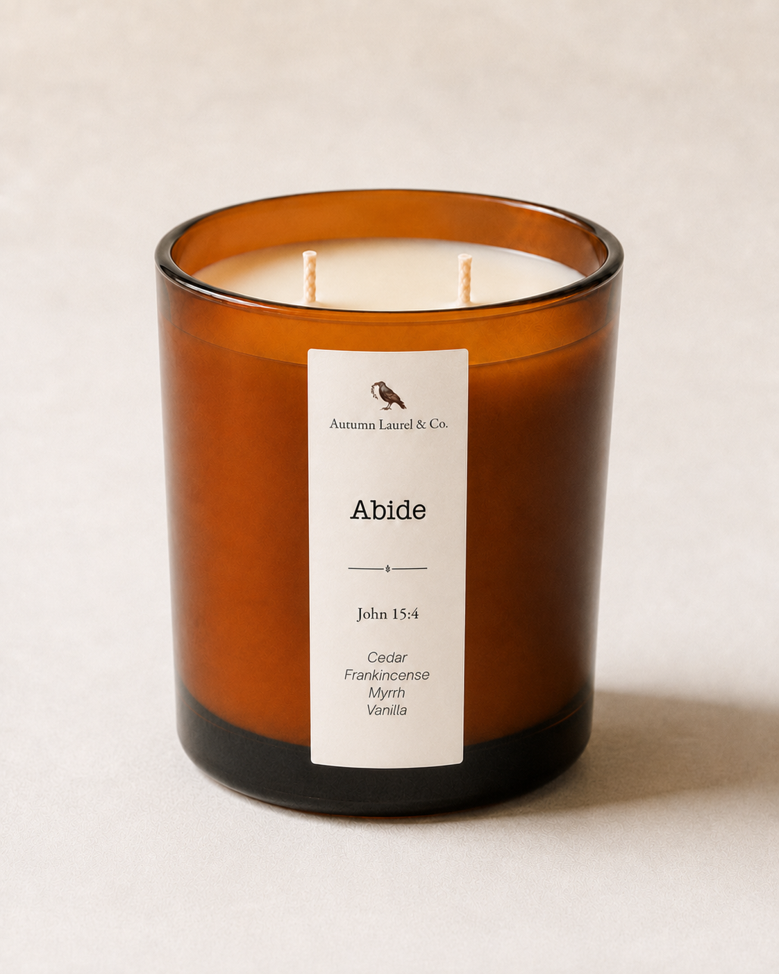 An amber glass jar candle with two wicks and a white label reading 'Abide' by Autumn Laurel & Co., photographed on a light neutral background.