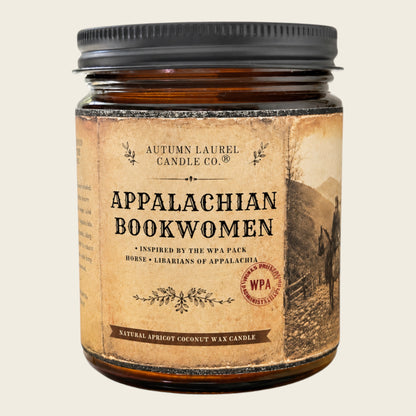 An amber glass jar candle with a black metal lid and a vintage-style tan label reading 'Appalachian Bookwomen' by Autumn Laurel Candle Co., shown on a light beige background.