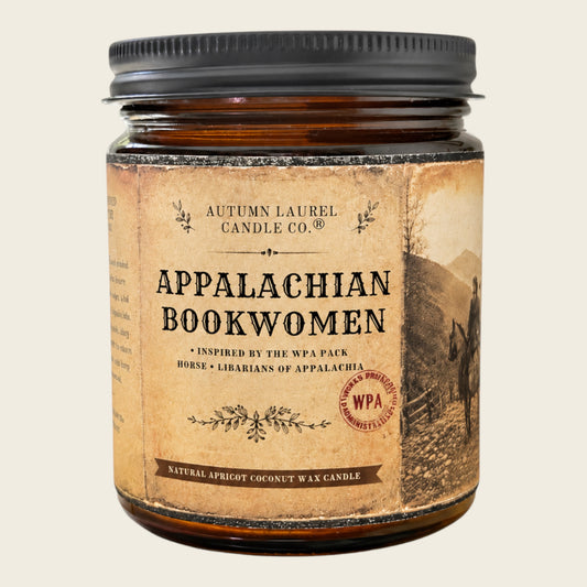 An amber glass jar candle with a black metal lid and a vintage-style tan label reading 'Appalachian Bookwomen' by Autumn Laurel Candle Co., shown on a light beige background.