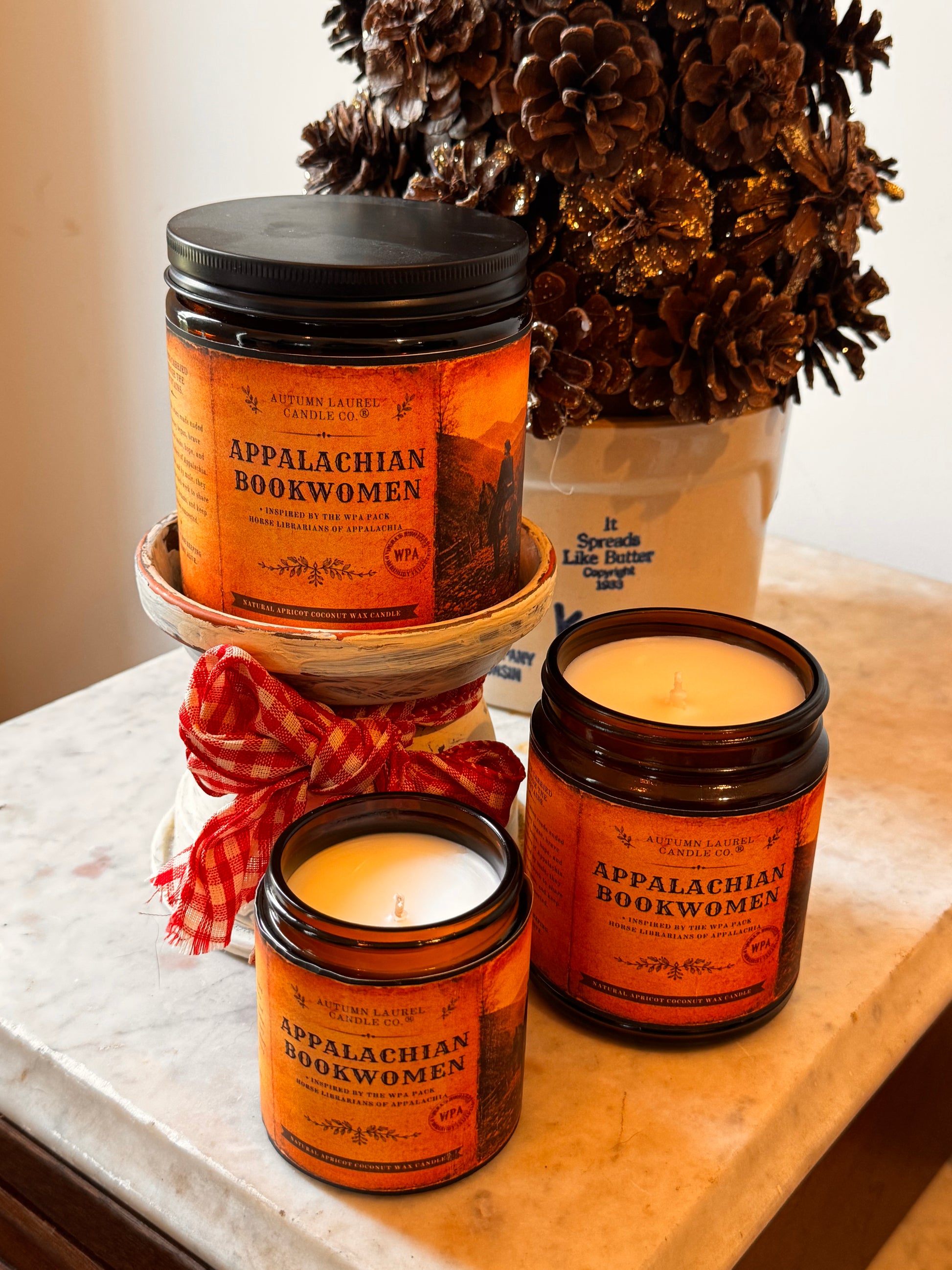 Three amber glass jar candles with orange labels reading Appalachian Bookwomen are arranged on a marble surface, with one candle elevated on a small stand and a red gingham ribbon around the middle jar. A white ceramic pot with pinecones sits in the background.
