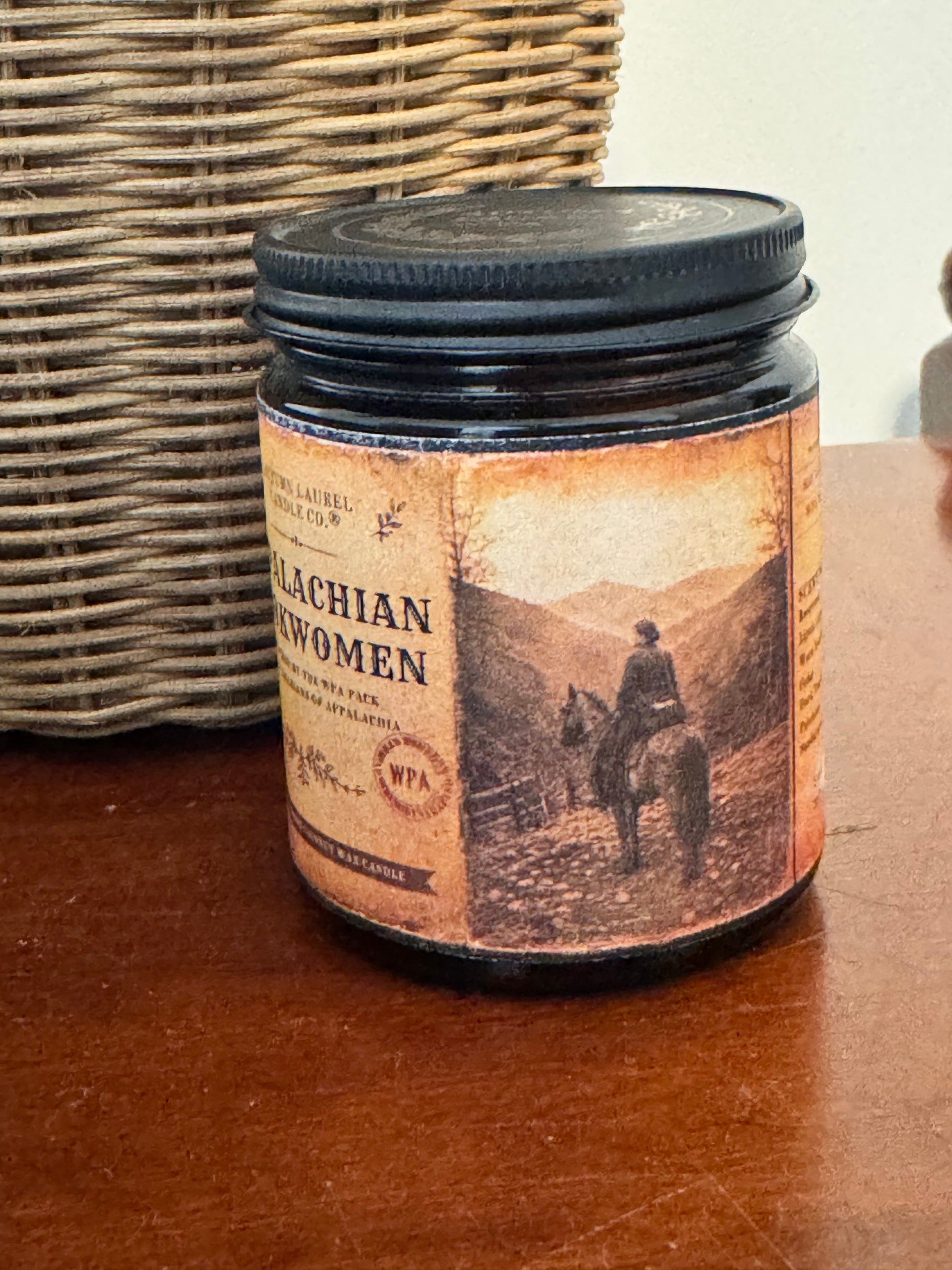 A sepia-toned archival illustration of an Appalachian woman riding horseback through the mountain hollers, inspired by the Pack Horse Librarians of the 1930s WPA era. The aged parchment-style label wraps around an amber glass candle jar with a matte black lid, creating the feel of a worn historical artifact.