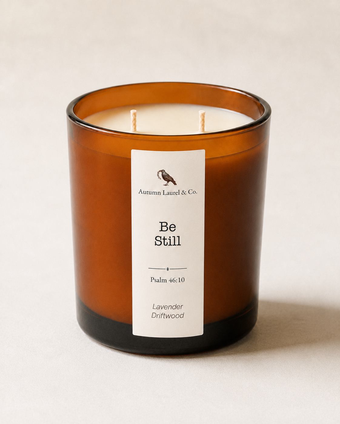 An amber glass jar candle with two wicks and a white label reading 'Autumn Laurel & Co. Be Still' on a light beige background.