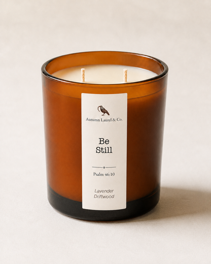 An amber glass jar candle with two wicks and a white label reading 'Autumn Laurel & Co. Be Still' on a light beige background.