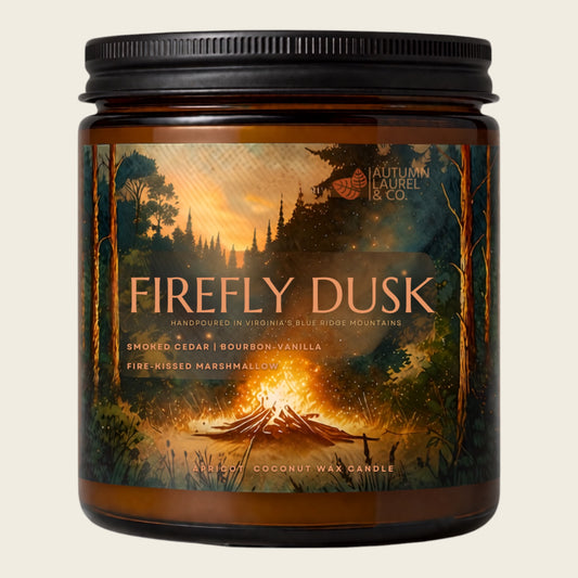 An amber glass jar candle with a black screw-top lid and a detailed label depicting a campfire scene at dusk. The label reads 'FIREFLY DUSK' and shows the brand 'Autumn Laurel & Co'.
