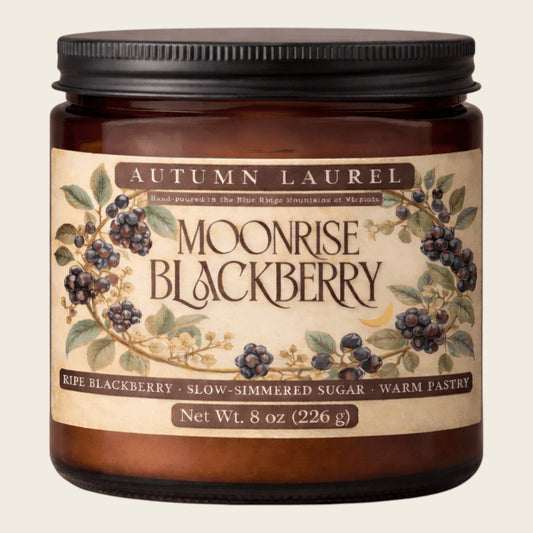 An 8 oz amber glass jar candle with a black metal lid and a vintage-style cream label reading 'Autumn Laurel Moonrise Blackberry'. The label features illustrations of blackberries and leaves on a light beige background.