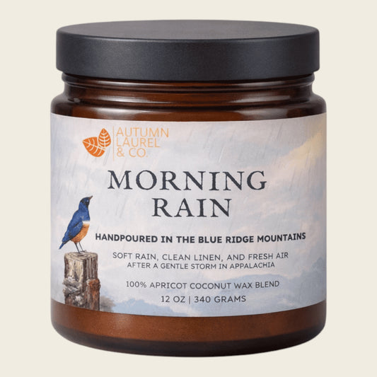 A 12 oz amber glass jar candle with a black lid and a white label reading 'MORNING RAIN' by Autumn Laurel & Co, featuring a bluebird illustration on a light cream background.
