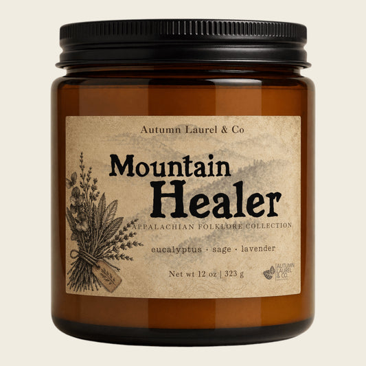 An amber glass jar candle with a black ribbed lid and a vintage-style beige label reading 'Mountain Healer' by Autumn Laurel & Co, shown on a light warm background.